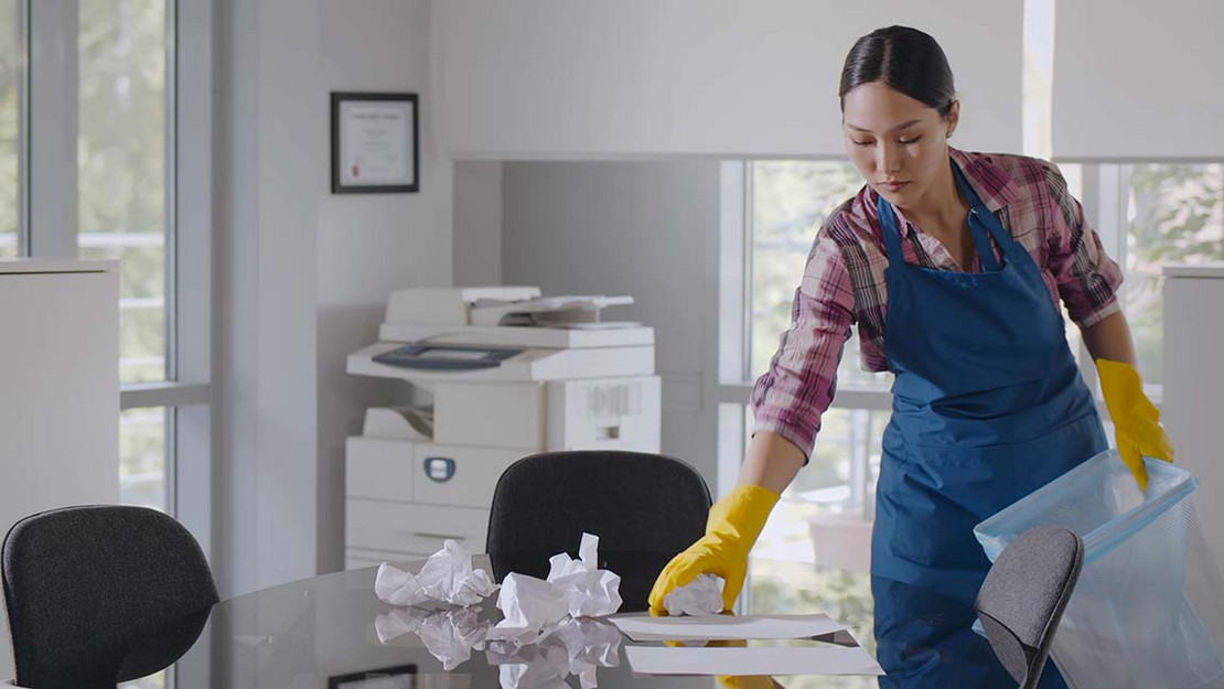 Frau in Putzkleidung räumt in einem Büro auf.
