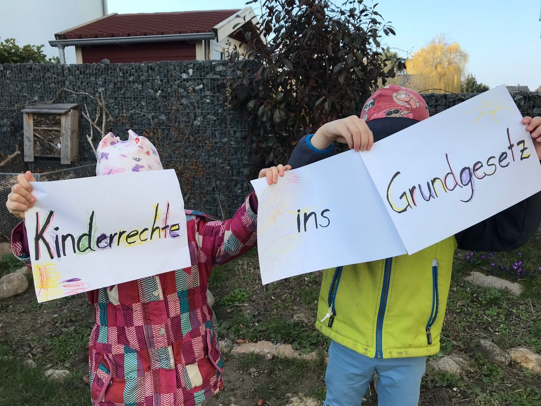 Zwei Kinder halten Zettel mit der Aufschrift "Kinderrecht ins Grundgesetz" hoch. 