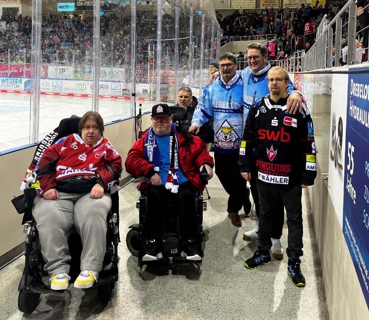 Personen mit Behinderung stehen bei einem Eishockeyspiel neben der Eisfläche. 