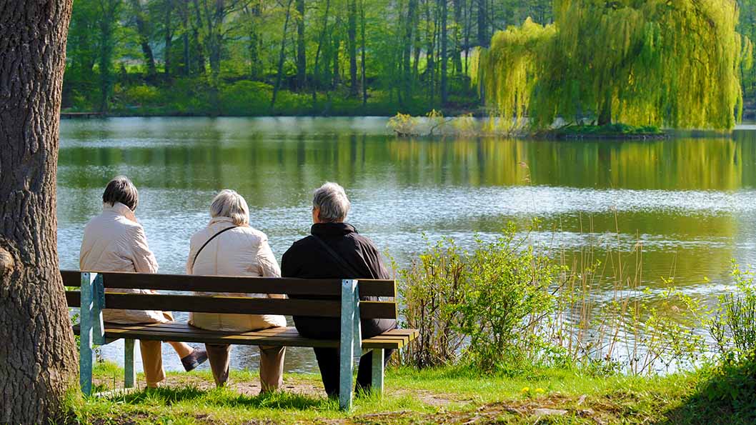 Drei Rentner sitzen bei sonnigem Wetter auf einer Bank an einem See. 